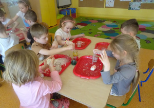 Dzieci barwią ciatolinę, pipetą nalewają barwniki na masę na tackach.